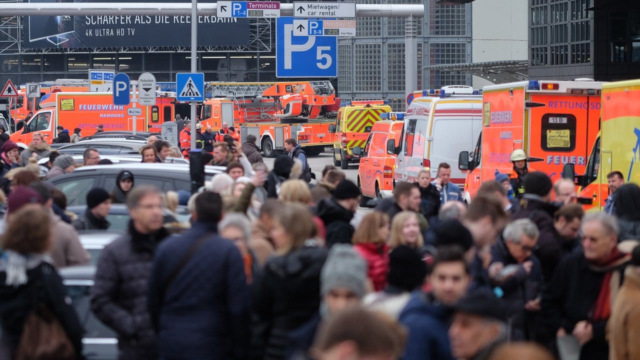 Reisegäste warten am Hamburger Flughafen zwischen Fahrzeugen der Feuerwehr auf Anweisungen.