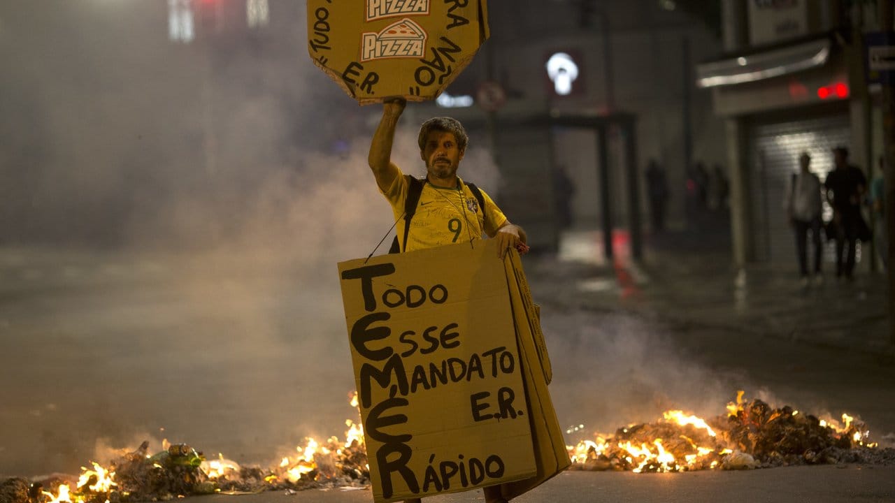 Vor einer brennenden Straßenblockade in Rio de Janeiro hält ein Mann Transparente gegen den Präsidenten.