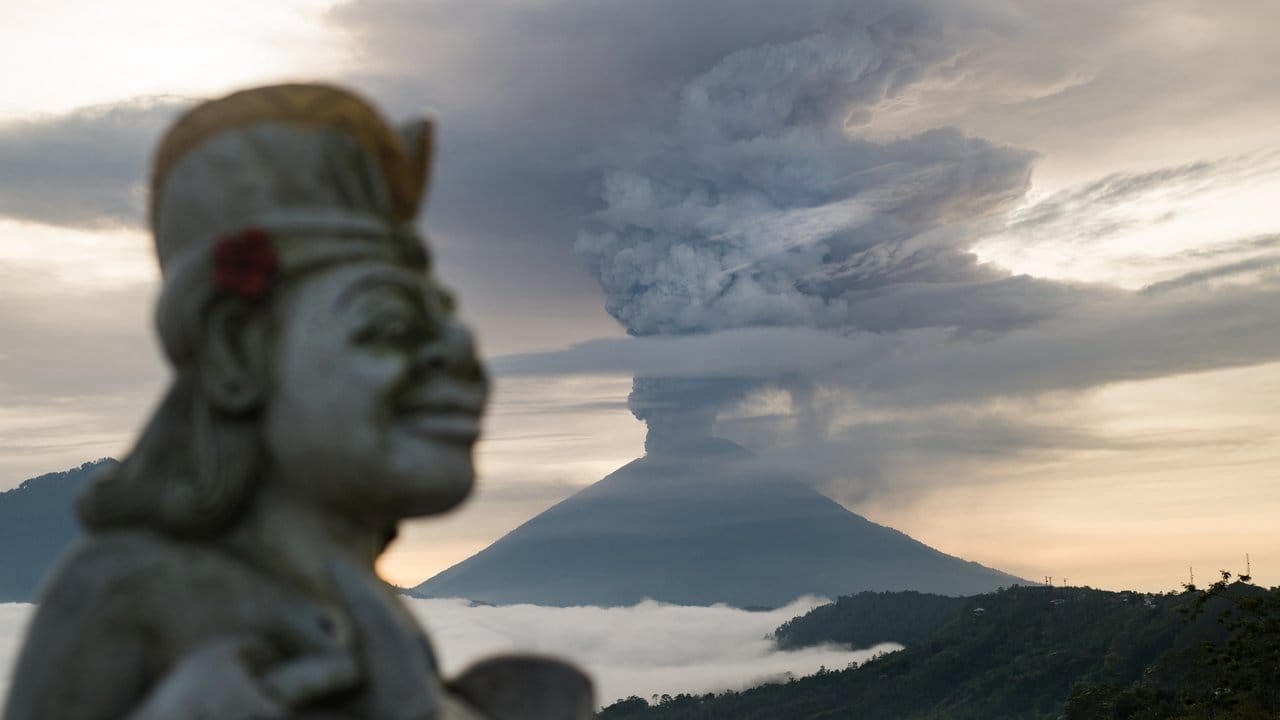 Entrücktes Lächeln: Eine Statue ist neben der riesigen Rauchsäule zu sehen, die über dem balinesischen Vulkan Mount Agung aufsteigt.