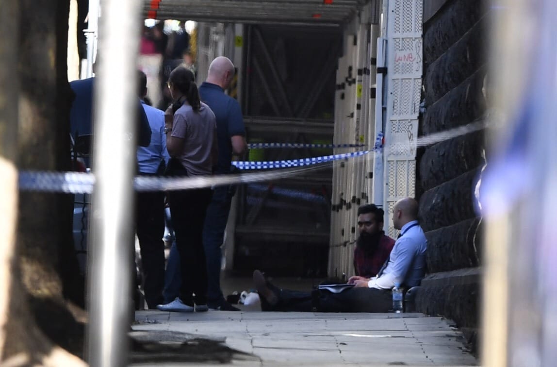 Zwei Männer sitzten nach dem Vorfall in Melbourne an eine Hauswand gelehnt auf einem Fußweg. Um sie herum stehen Polizisten.