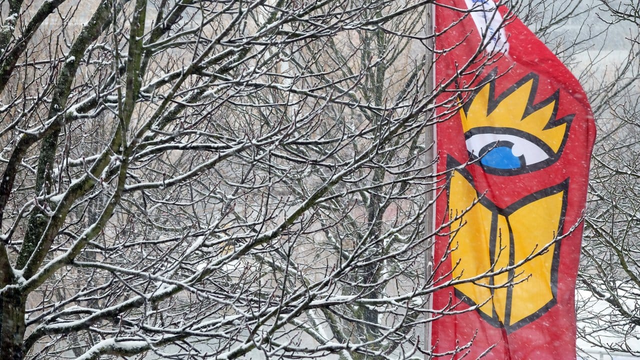 Eine Fahne mit dem Logo der Leipziger Buchmesse weht vor verschneiten Bäumen am Messegelände.