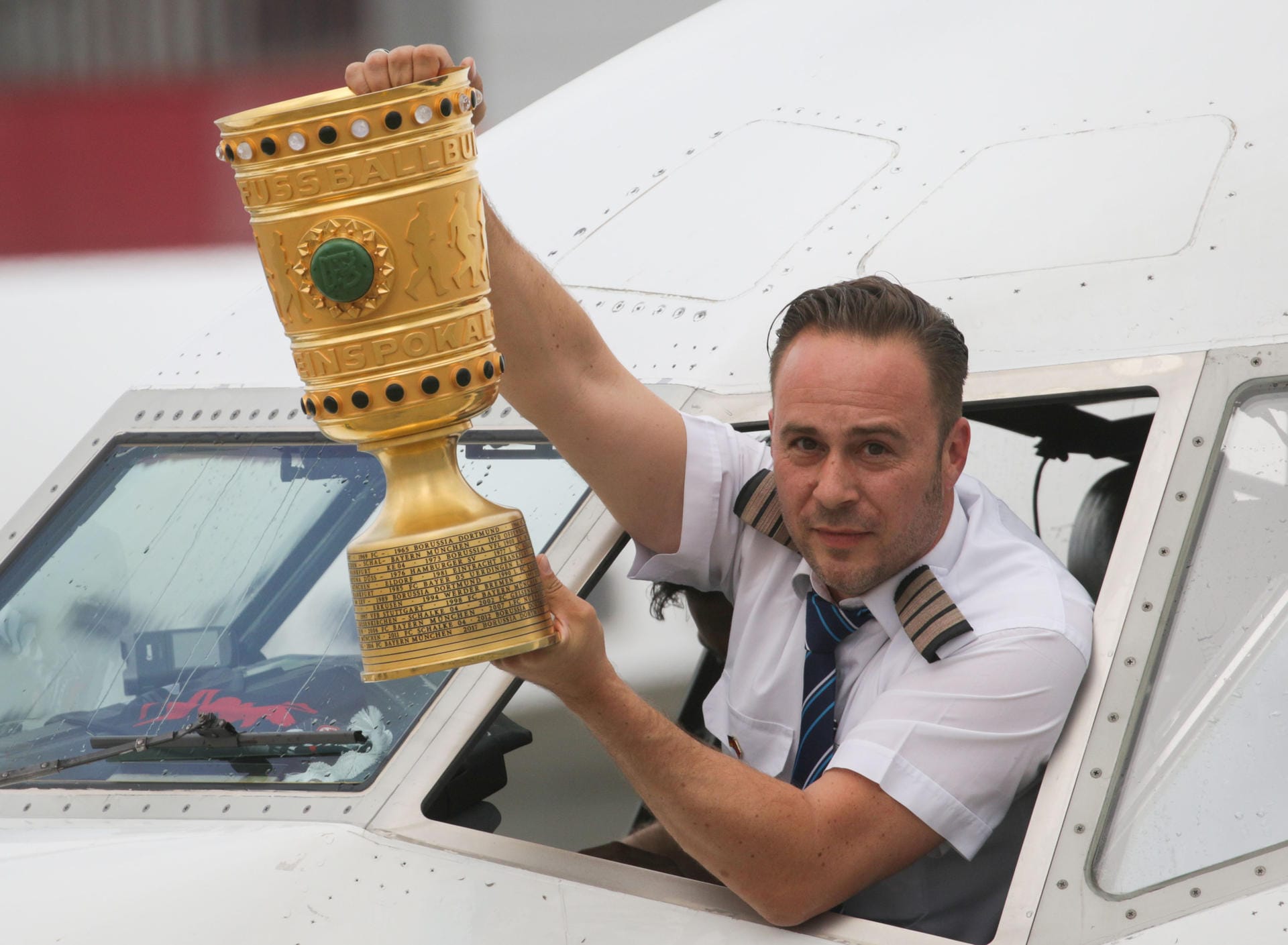 Flugkapitän Robert Weller hält nach der Landung des Flugzeugs mit den Spielern von Eintracht Frankfurt den DFB-Pokal aus dem Cockpitfenster.