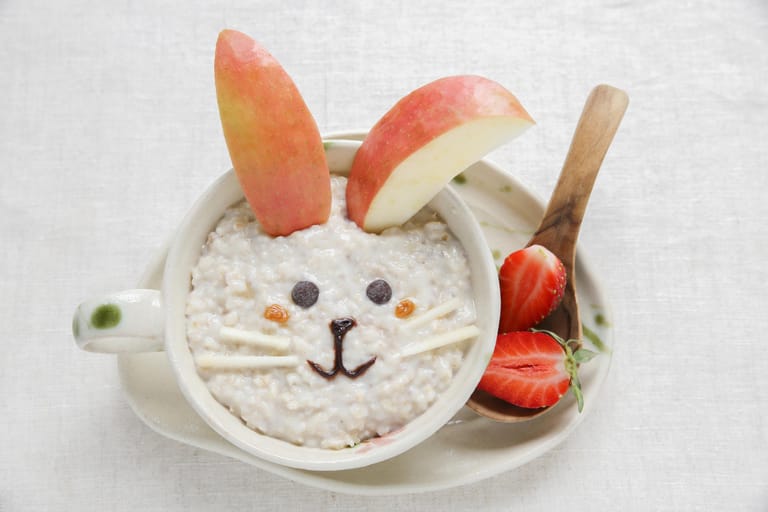 Haferflocken-Frühstück für Kinder: Peppen Sie den Brei lustig auf – mit Apfelschnitzohren, zwei Rosinen oder Beeren als Augen, Schnurrhaaren aus geschnittenen Apfelstreifen und Schokolade als Näschen.