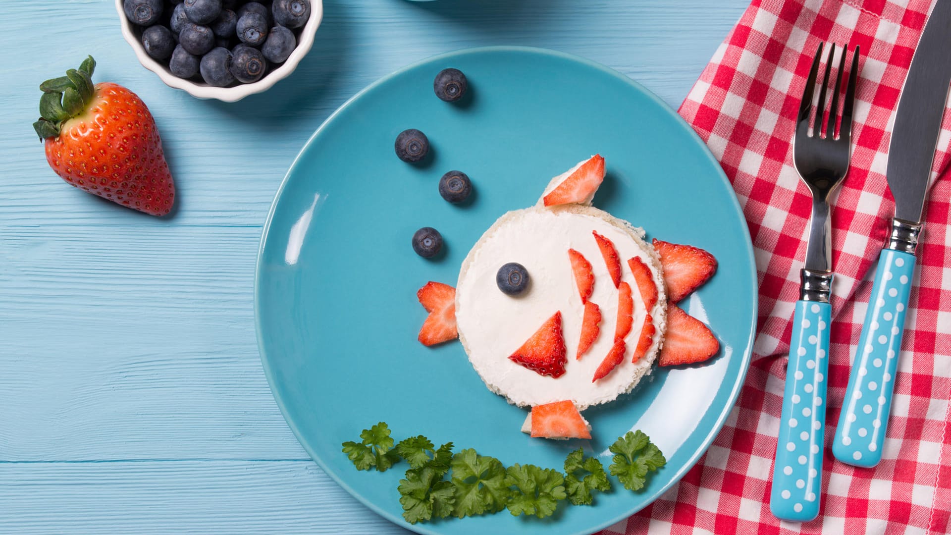 Lustiger Toast in Fischform: Bestreichen Sie einen runden Toast mit Frischkäse. Belegen Sie diesen mit geschnittenen Erdbeeren und einer Heidelbeere und dekorieren Sie den Teller mit den restlichen Heidelbeeren sowie Petersilie.