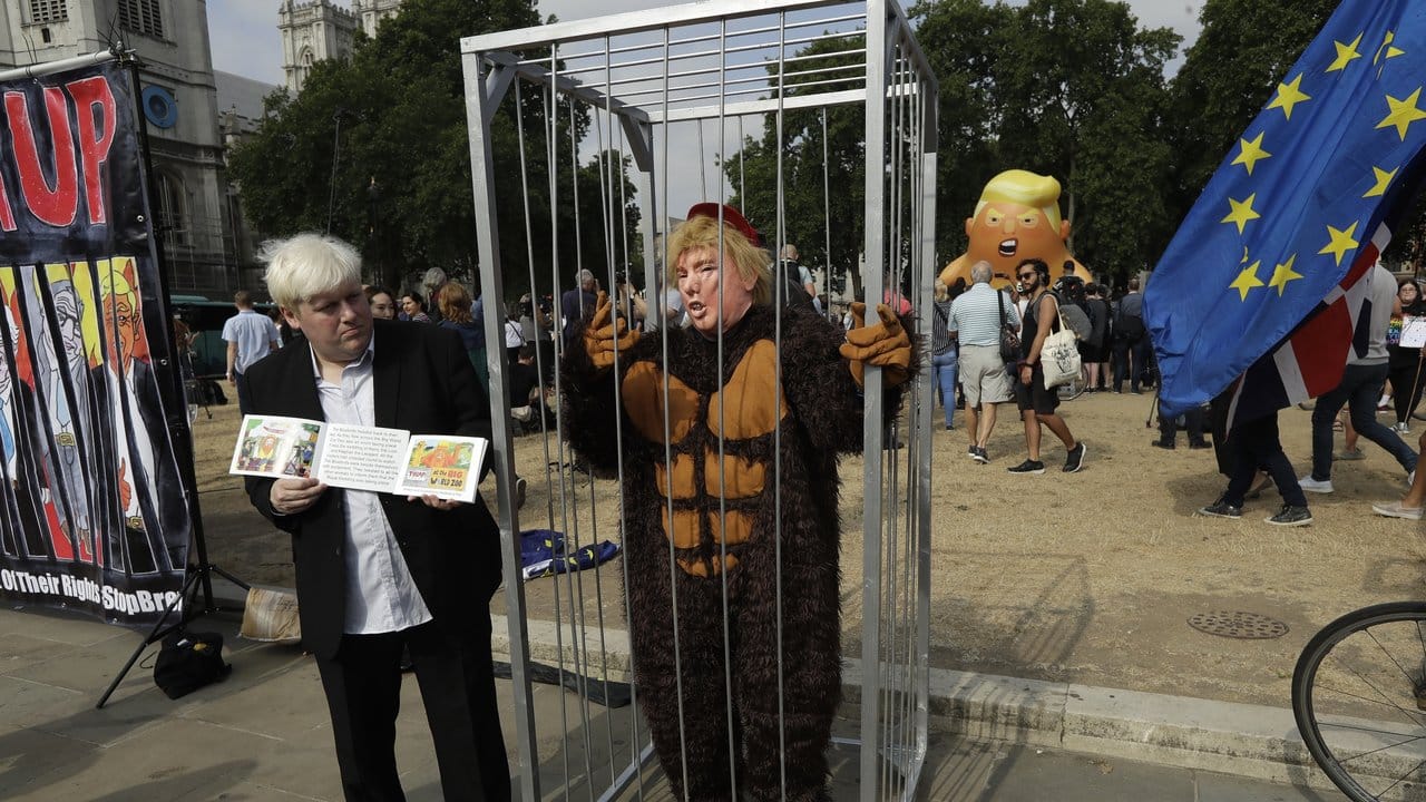 Ein Boris Johnson-Double steht neben einem Käfig, in dem ein Mann im Affenkostüm und Trump-Maske steht.