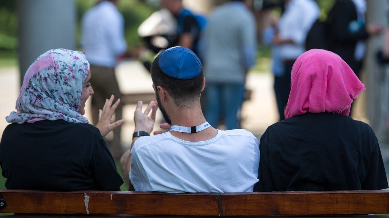 Zwei junge Muslimas mit Kopftuch und ein junger jüdischer Mann mit Kippa unterhalten sich miteinander.