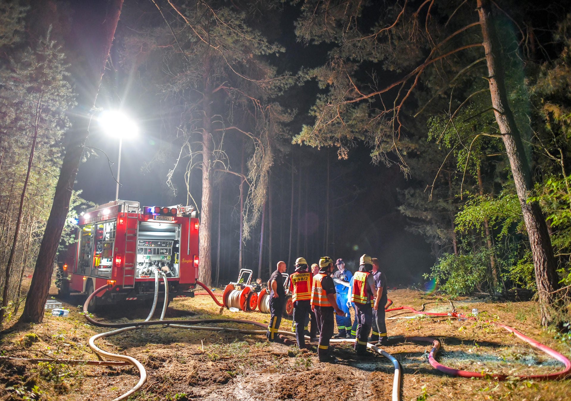 Einsatzkräfte der Feuerwehr stehen an einer Pumpstation für den Wassertransport. Das Feuer hat sich auf einer großen Waldfläche ausgebreitet.