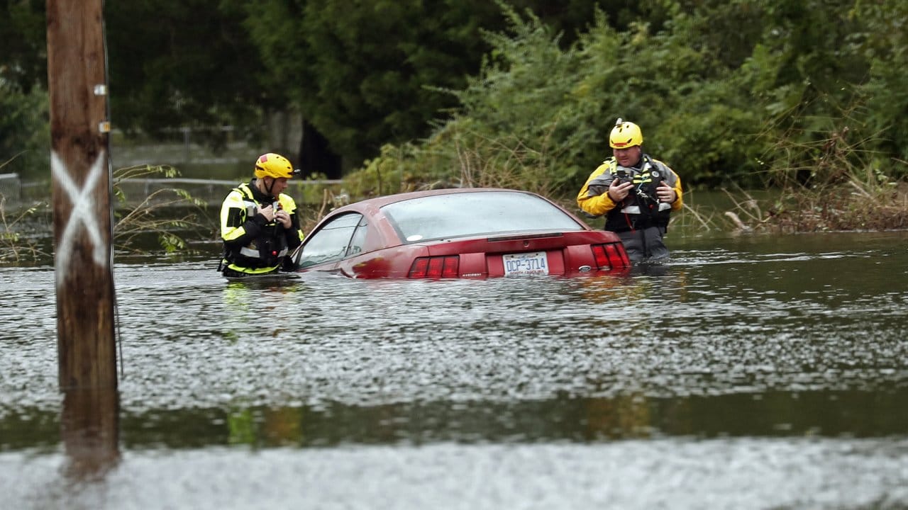 Mitglieder der Feuerwehr untersuchen auf einer überfluteten Straße ein halb unter Wasser stehendes Auto.