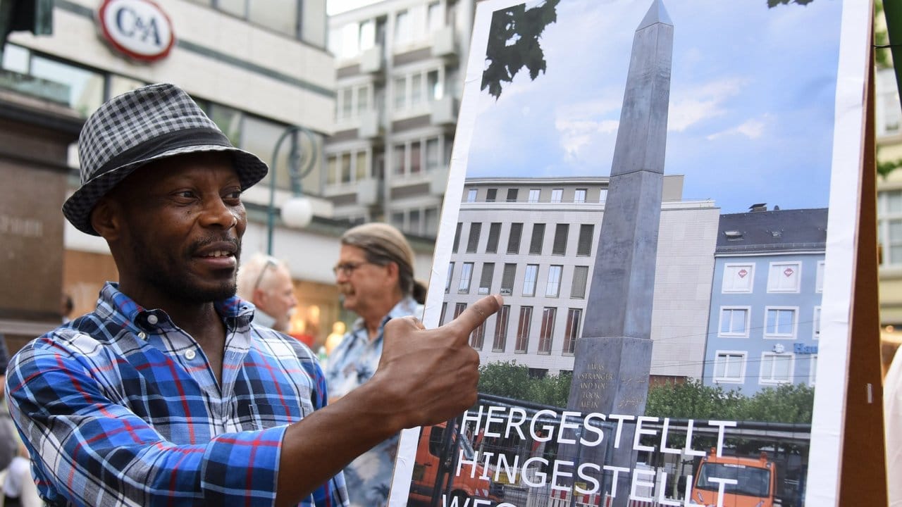 Der documenta-Künstler Olu Oguibe steht neben einem Plakat, das seinen Obelisken zeigt.
