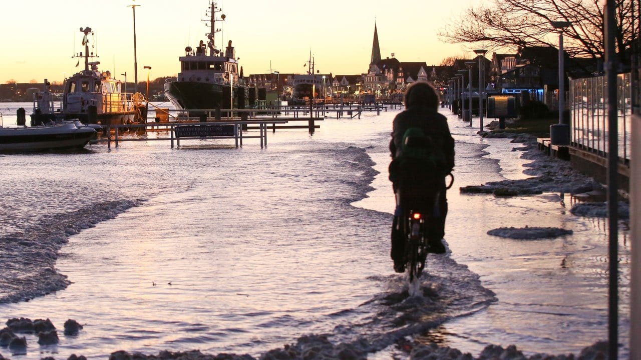 Ein Radfahrer fährt mit seinem Fahrrad die durch Hochwasser überflutete Strandpromenade von Lübeck-Travemünde entlang.