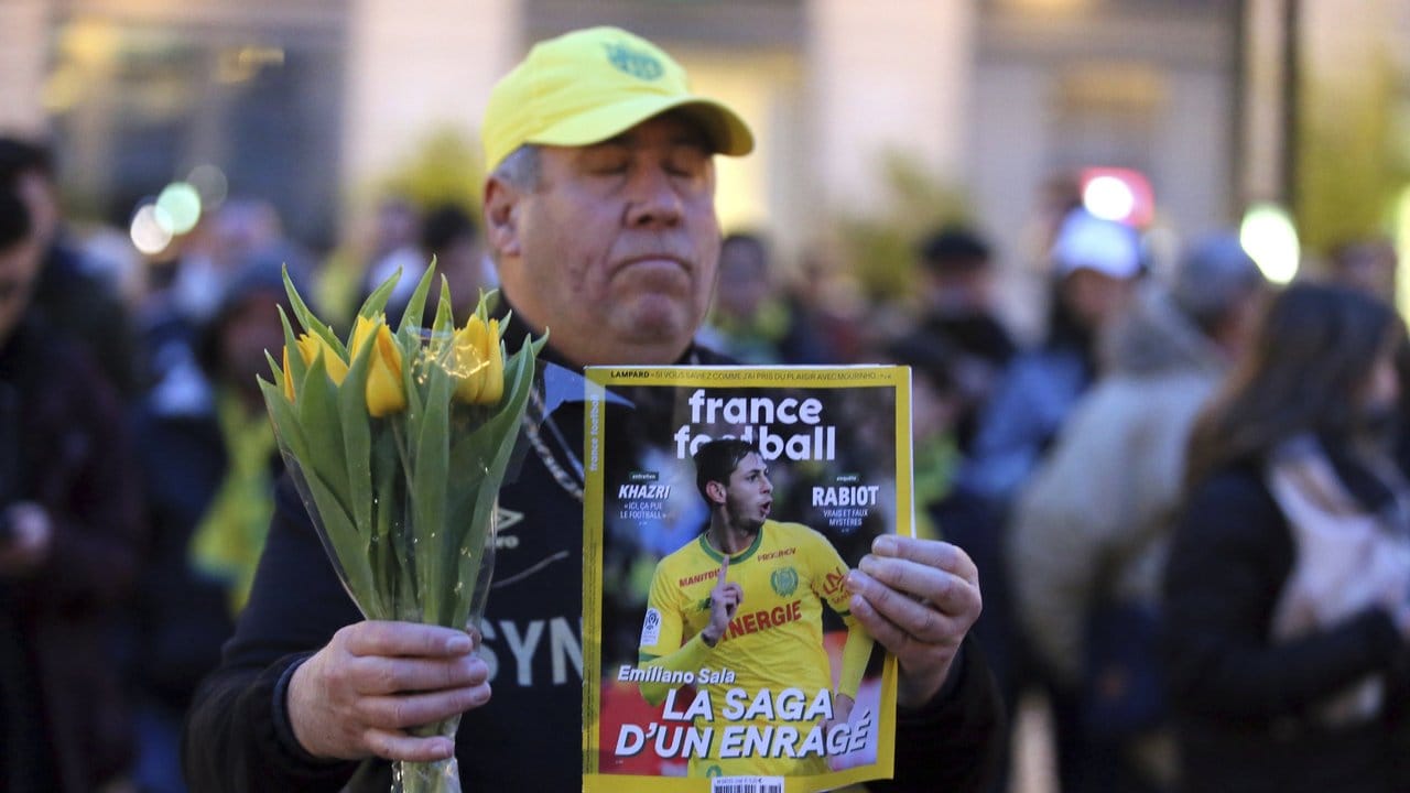 Ein Fan des FC Nantes hält gelbe Blumen und ein Fußballmagazin mit dem verschollenen Fußballspieler Emiliano Sala auf dem Cover.