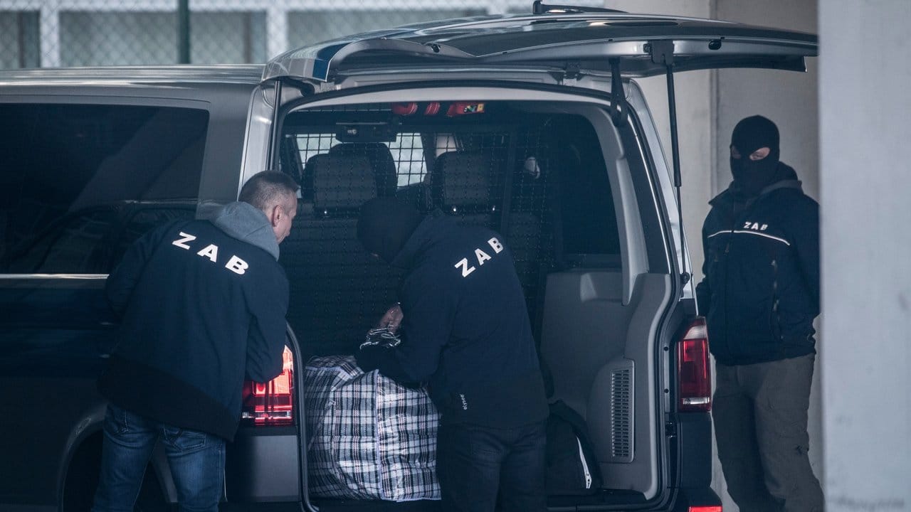 Mit Sturmhauben maskierte Mitarbeiter der Zentralen Ausländerbehörde entladen das Gepäck eines Abschiebehäftlings.