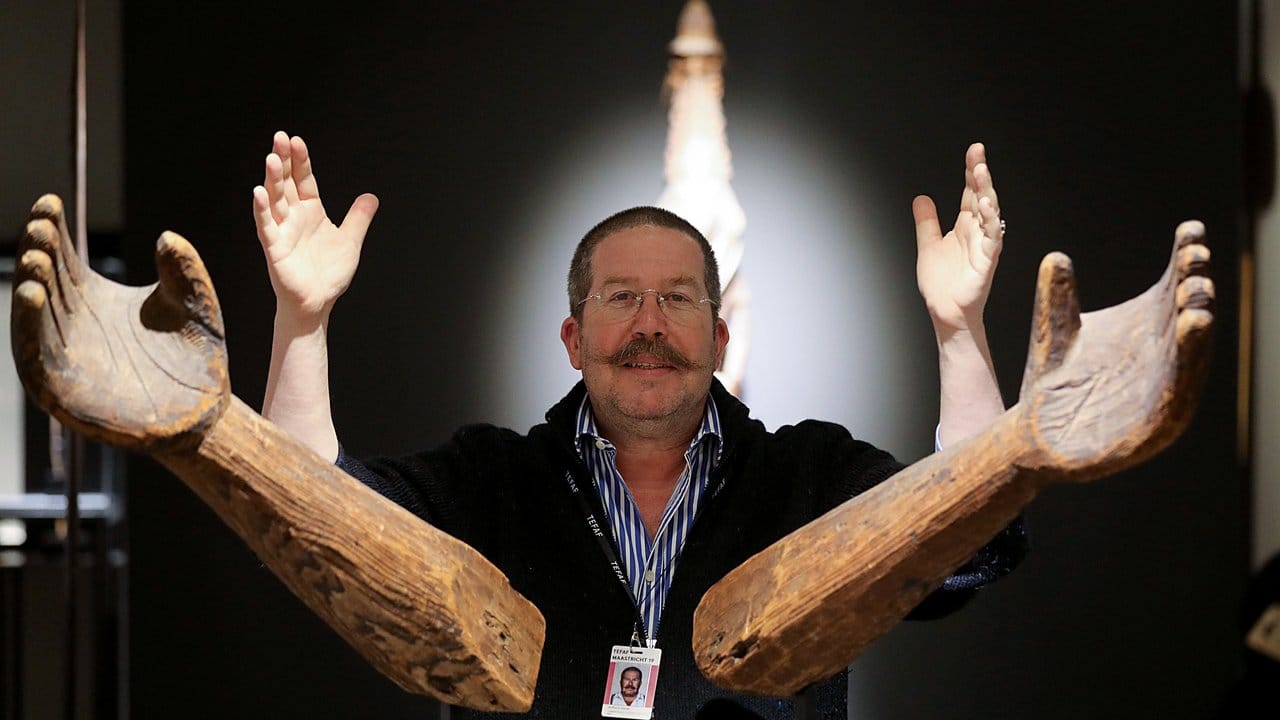 Galerist Anthony Meyer auf dem Stand seiner Galerie auf der Kunstmesse TEFAF hinter Unterarmen aus Holz eines Indianerstammes aus British Columbia.