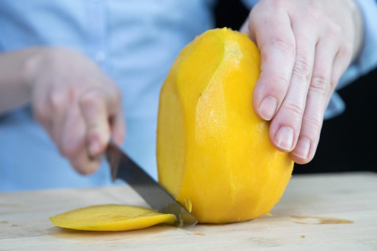 Mango: Schritt 2 – Die geschälte Frucht aufrecht auf das Schneidbrett stellen und gut festhalten. Dann mit einem mittelgroßen Messer am Kern vorbei eine etwa drei Millimeter dicke Scheibe abschneiden.