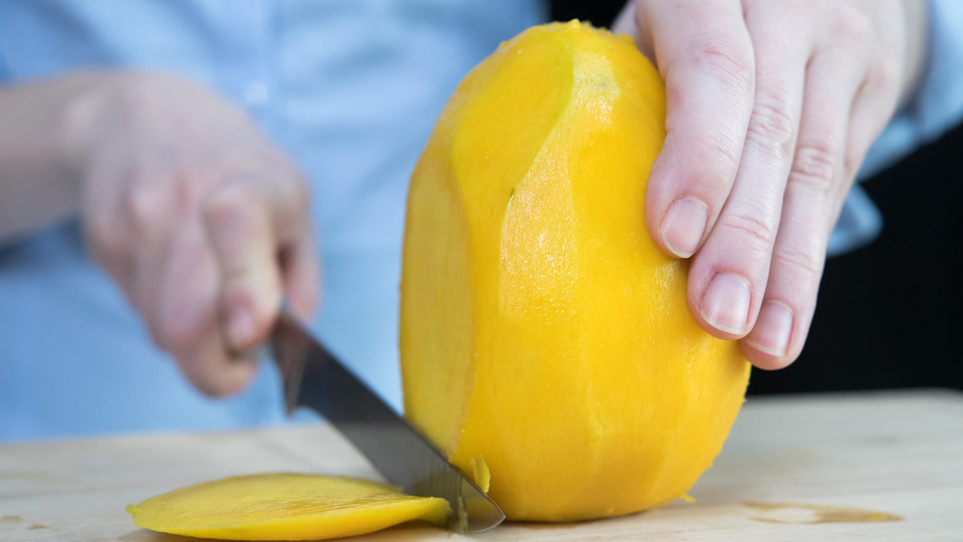 Mango: Schritt 2 – Die geschälte Frucht aufrecht auf das Schneidbrett stellen und gut festhalten. Dann mit einem mittelgroßen Messer am Kern vorbei eine etwa drei Millimeter dicke Scheibe abschneiden.