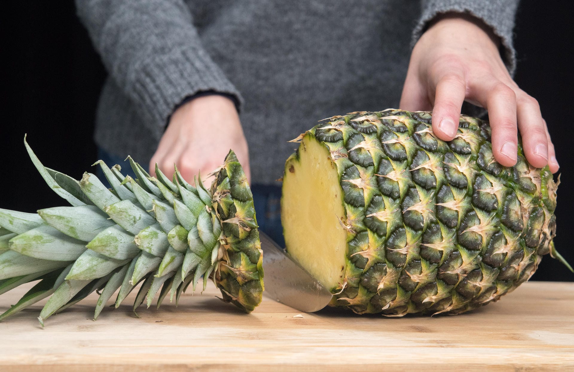 Ananas richtig aufschneiden: Schritt 1 – Ananas quer vor sich aufs Brett legen und mit einem mittelgroßen Messer den Blütenansatz abschneiden.
