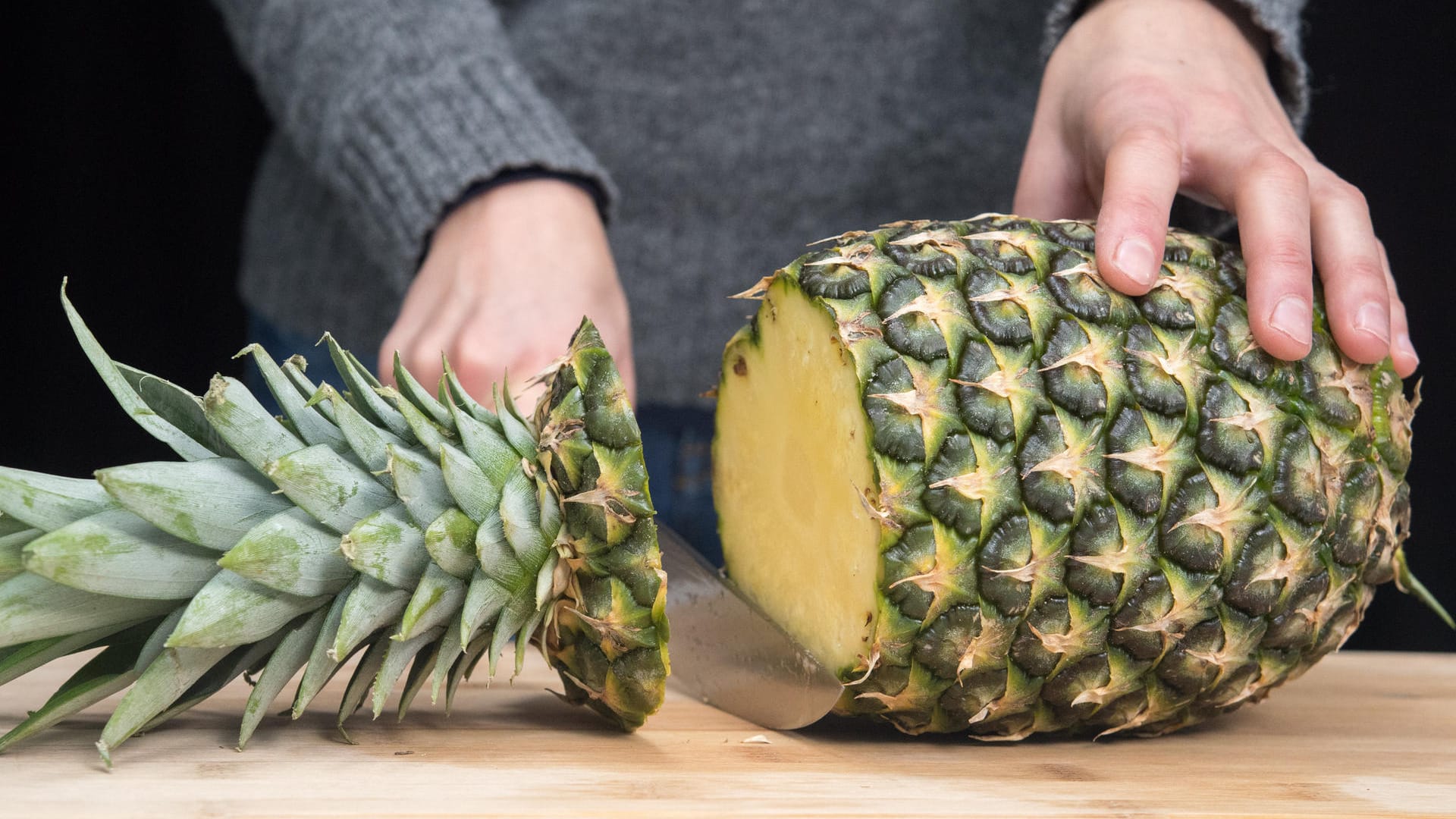 Ananas richtig aufschneiden: Schritt 1 – Ananas quer vor sich aufs Brett legen und mit einem mittelgroßen Messer den Blütenansatz abschneiden.