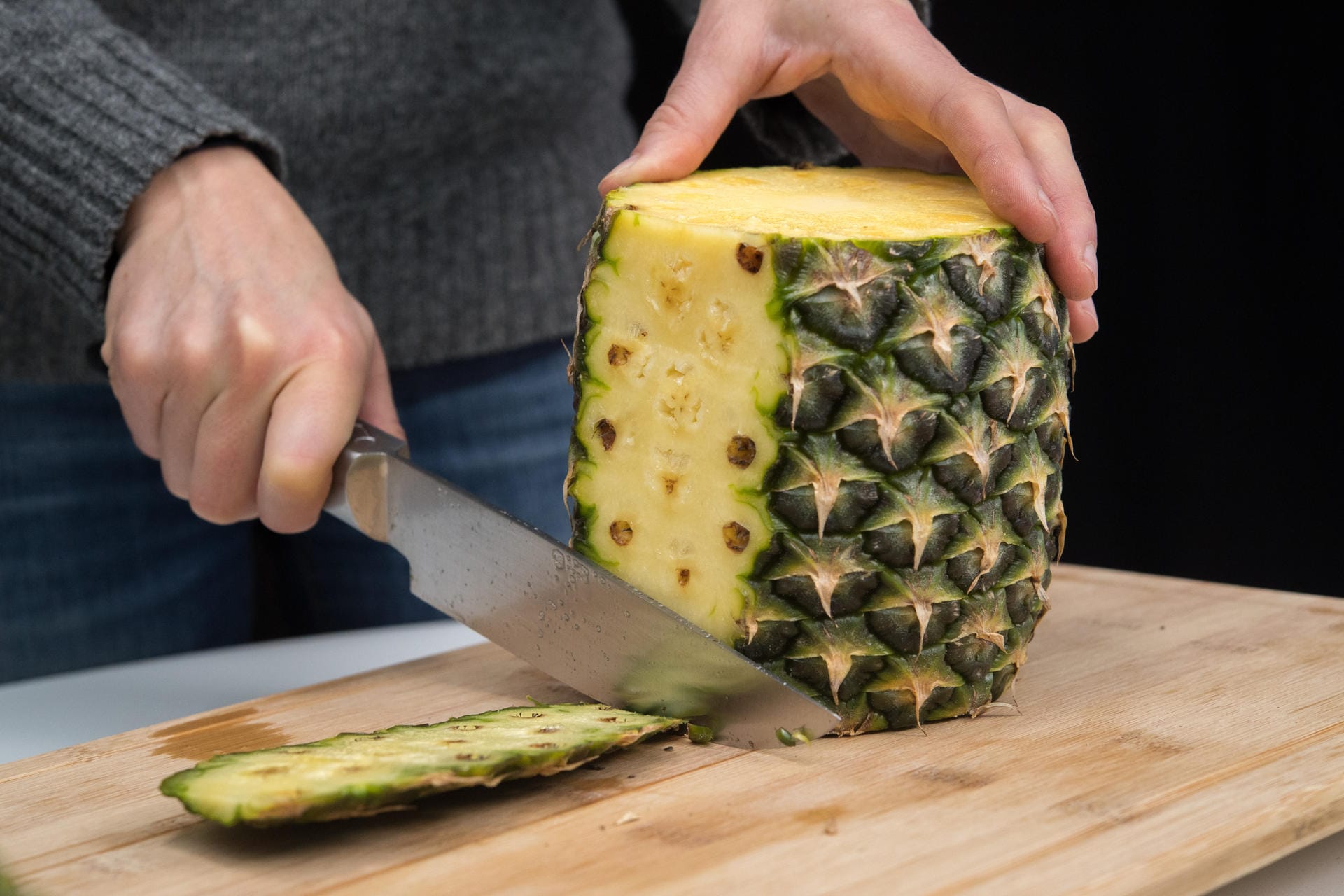 Ananas richtig aufschneiden: Schritt 3 – Die Frucht aufrecht auf das Brett stellen. Dann mit einem Messer der Wölbung folgend die Schale von oben nach unten abschneiden.