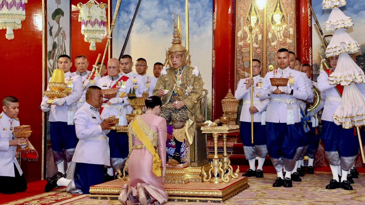Thailands König Maha Vajiralongkorn (M) auf dem Thron, vor ihm kniet seine Frau, Königin Suthida.