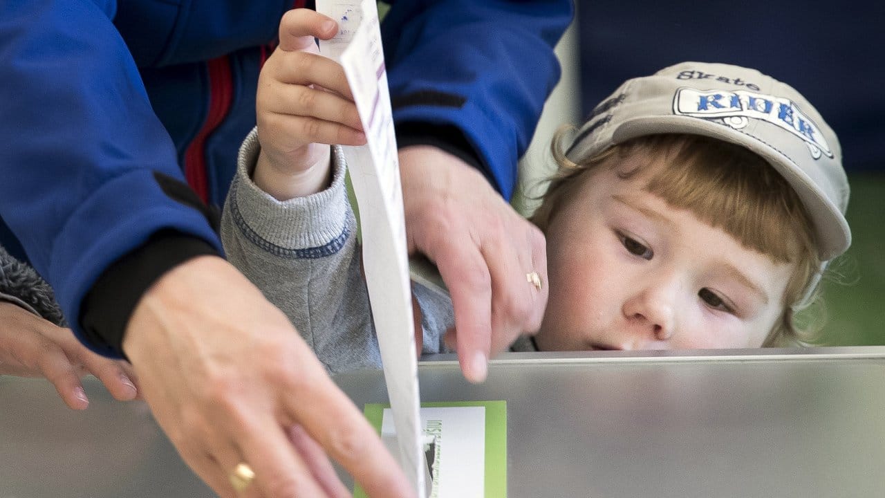 Eine Frau lässt ein Kind ihren Stimmzettel in die Wahlurne stecken.