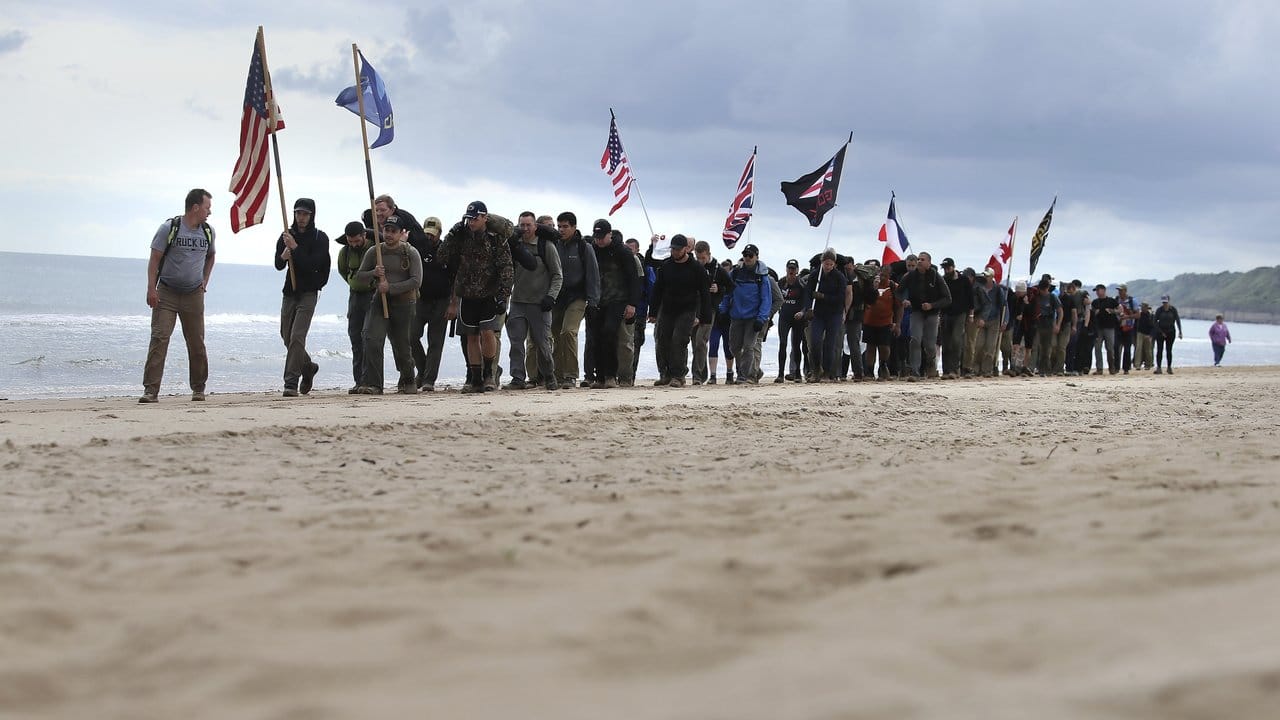 Männer gehen mit Flaggen der alliierten Truppen den Omaha-Beach in der Normandie entlang.