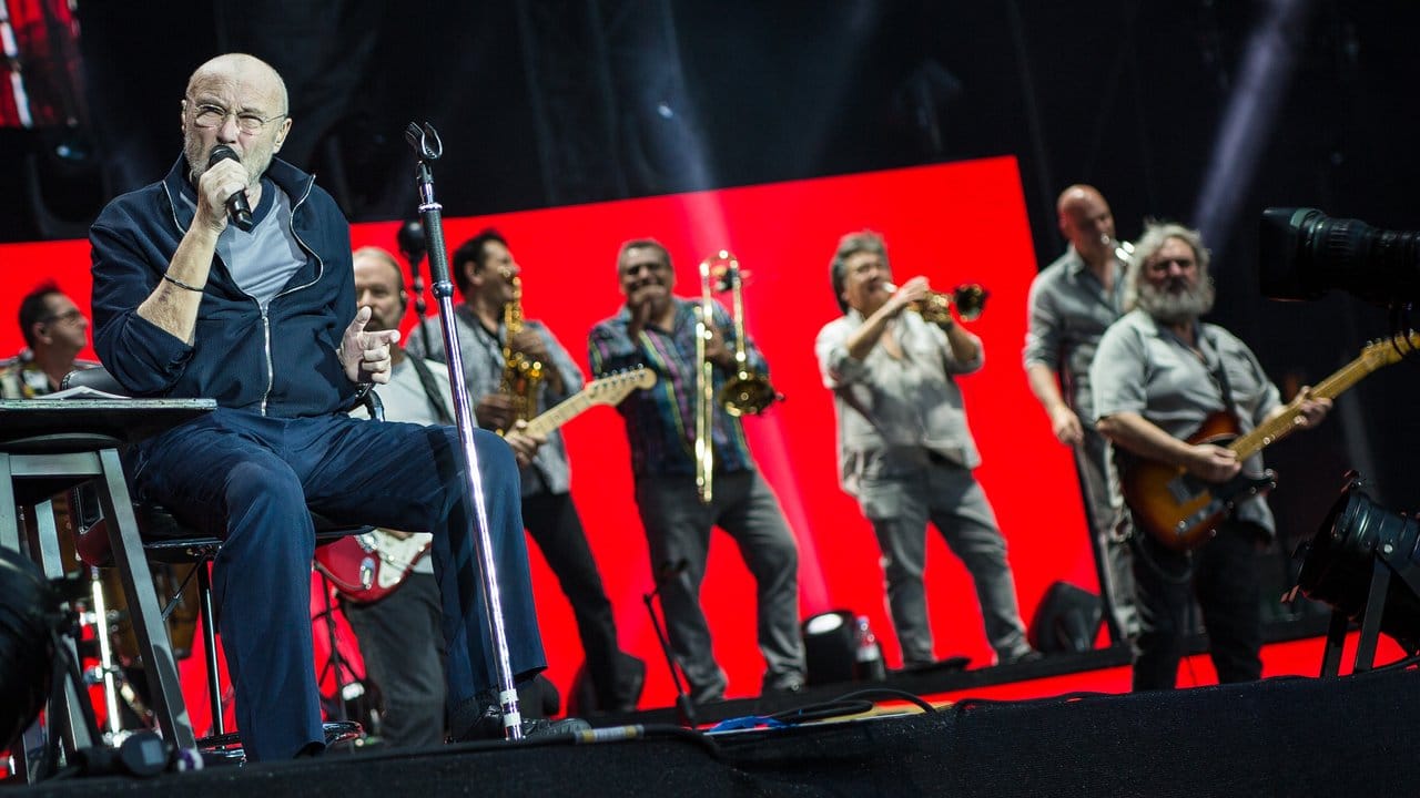 Sänger Phil Collins sitzt auf der Bühne und singt, hinter ihm stehen die Bandmitglieder.