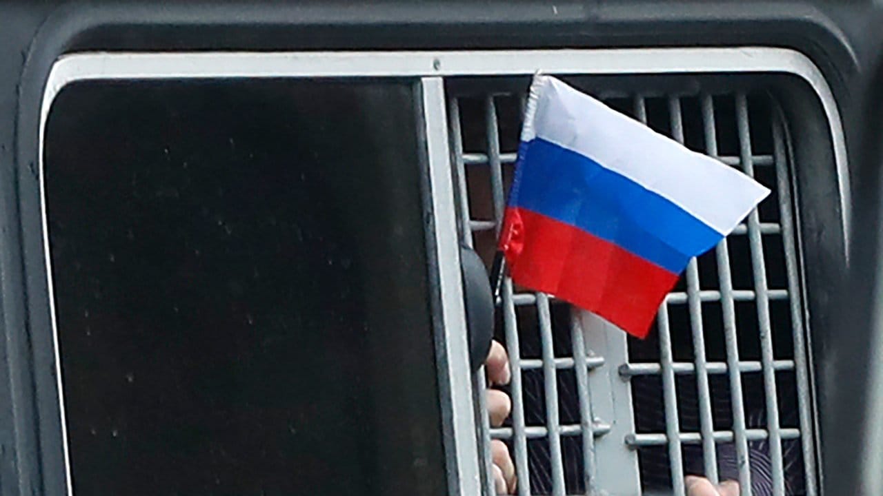 Ein festgenommener Demonstrant schwenkt aus einem Polizeiwagen heraus eine kleine russische Fahne.