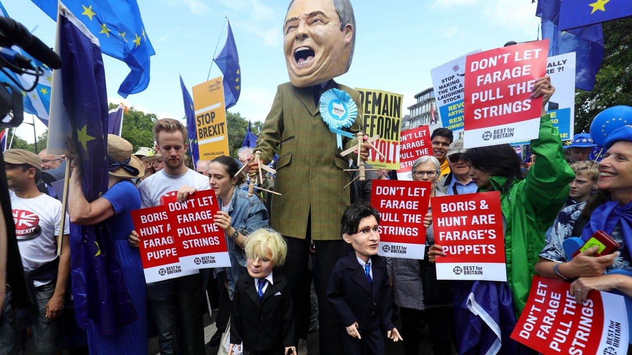Ein Demonstrant (M) hat sich für den Protestzug als Nigel Farage, Vorsitzender der Brexit-Partei, verkleidet - und hält die Schnüre der Marionetten "Boris Johnson" und "Jeremy Hunt" in den Händen.