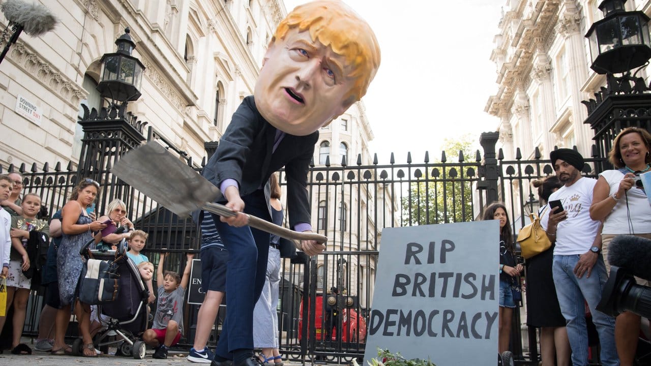 Ein Mann verkleidet mit einem riesigen Kopf des britischen Premierministers Johnson neben einem symbolischen Grabstein mit der Aufschrift "RIP British Democracy" (Ruhe in Frieden britische Demokratie).