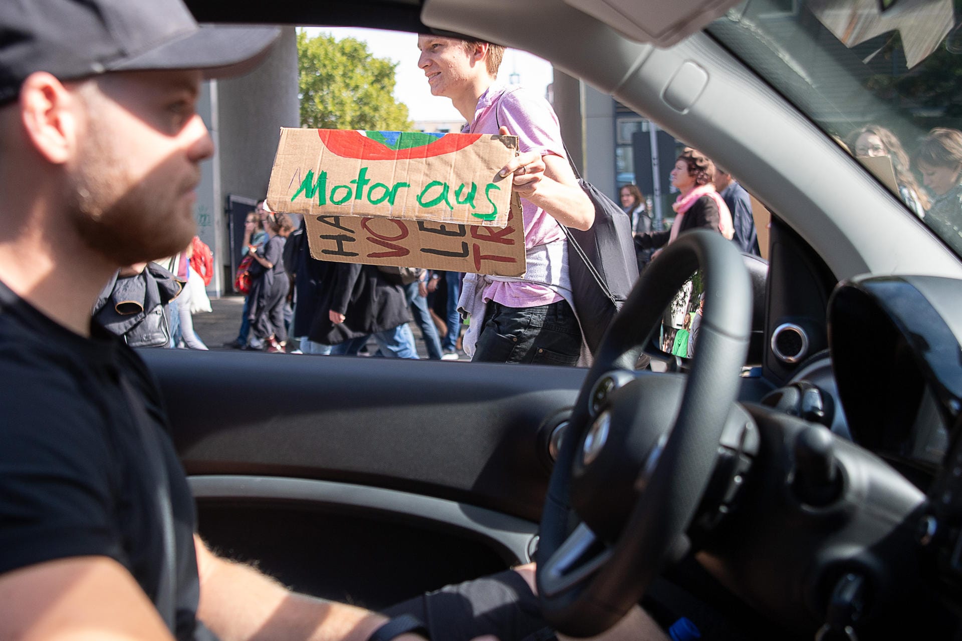 In Stuttgart wartet ein Mann in seinem Auto, das auf einer blockierten Straße steht. Neben ihm steht ein Demonstrant und hält ein Schild mit der Aufschrift "Motor aus" hoch.