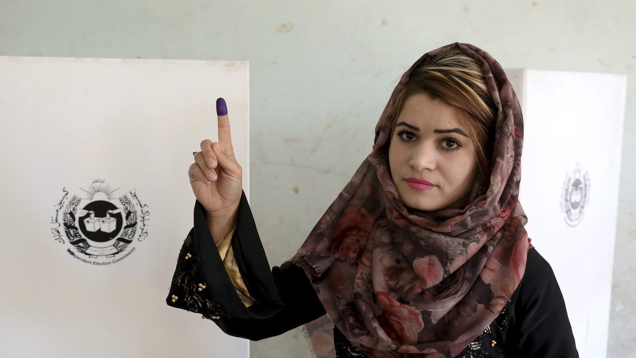 Eine Wählerin zeigt ihren mit Wahltinte markierten Zeigefinger vor einer Wahlkabine.