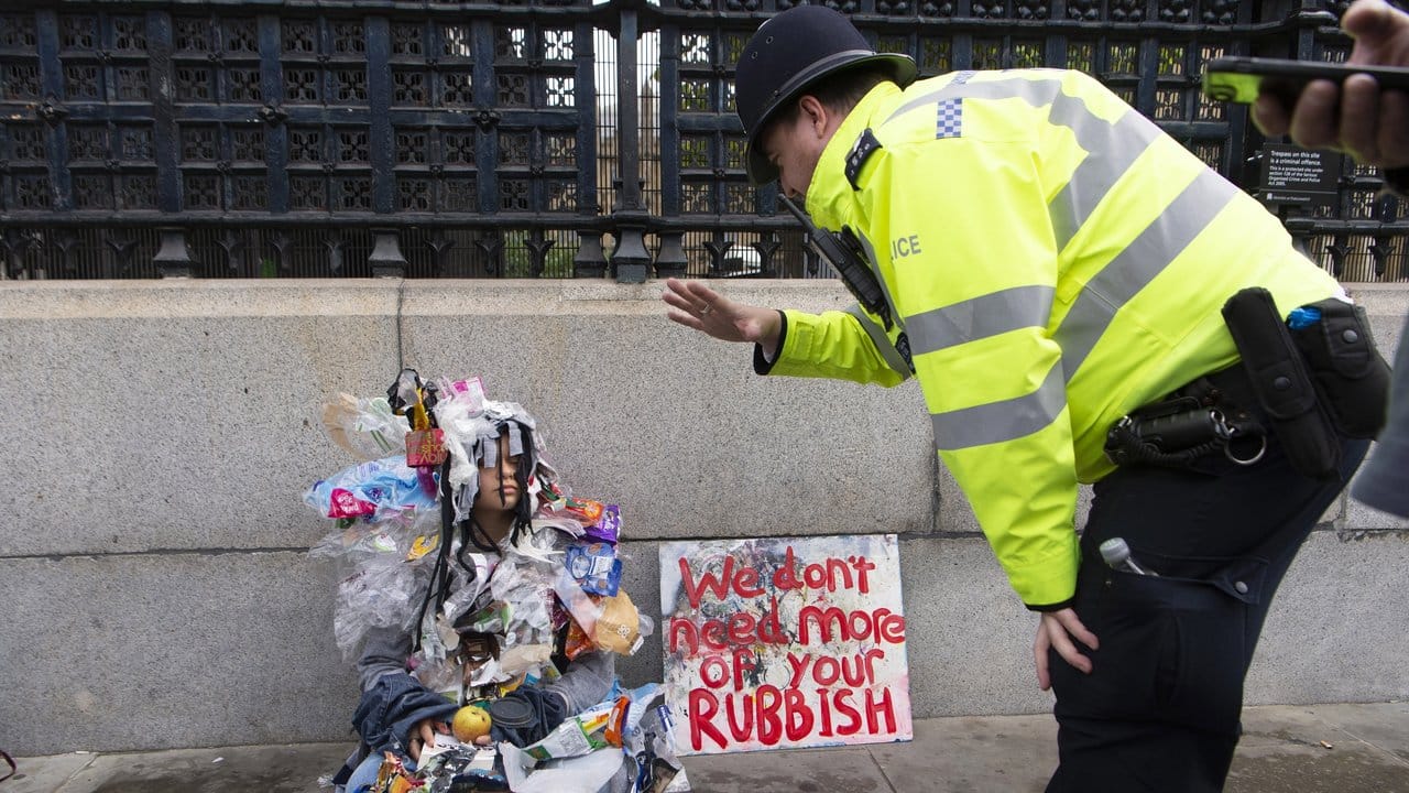 Ein Polizist spricht in London mit einer mit Müll bedeckten Klima-Aktivistin, die neben einem Plakat mit der Aufschrift "We don't need more of your rubbish" (Wir brauchen nicht mehr von eurem Müll) sitzt.