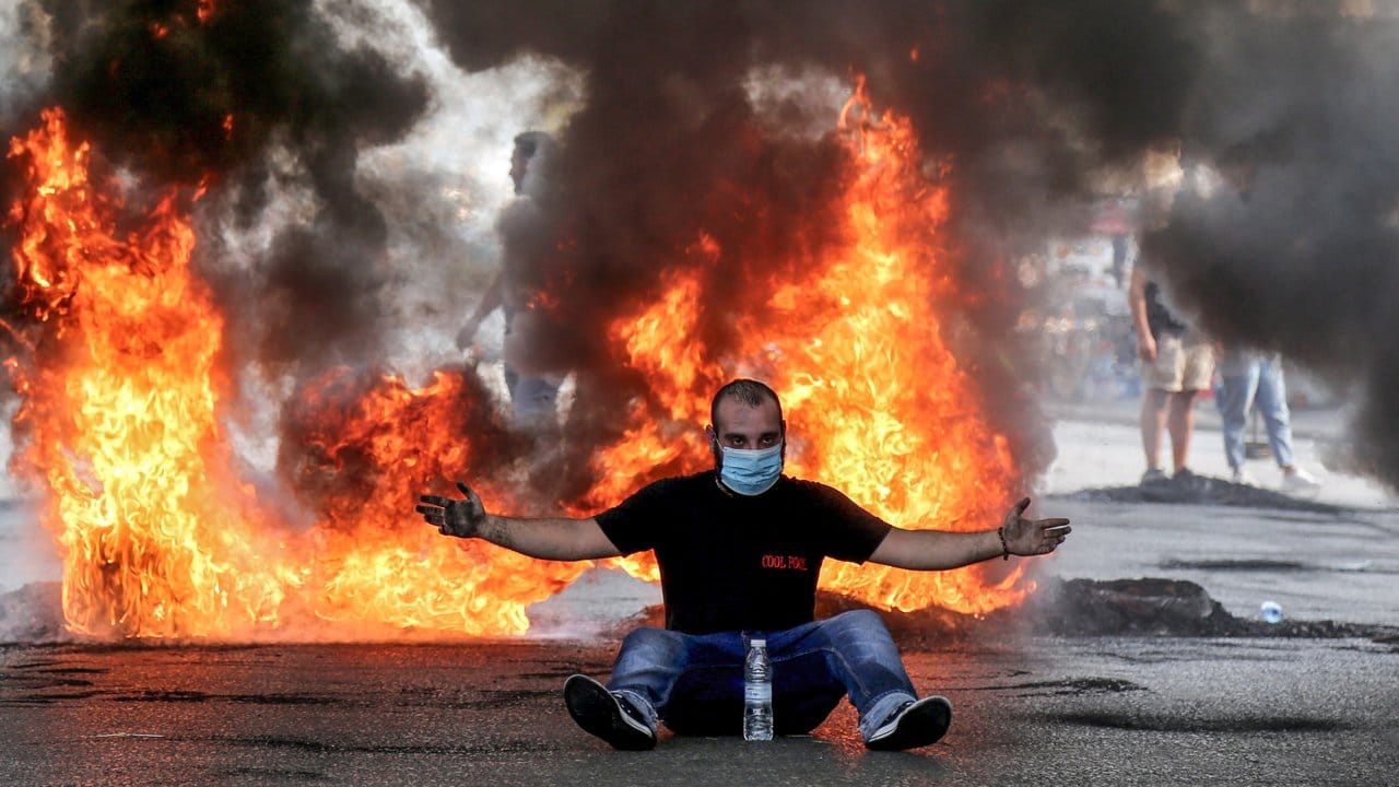 Ein Demonstrant mit Mundschutz sitzt mit ausgestreckten Armen vor einer brennenden Straßenbarrikade.