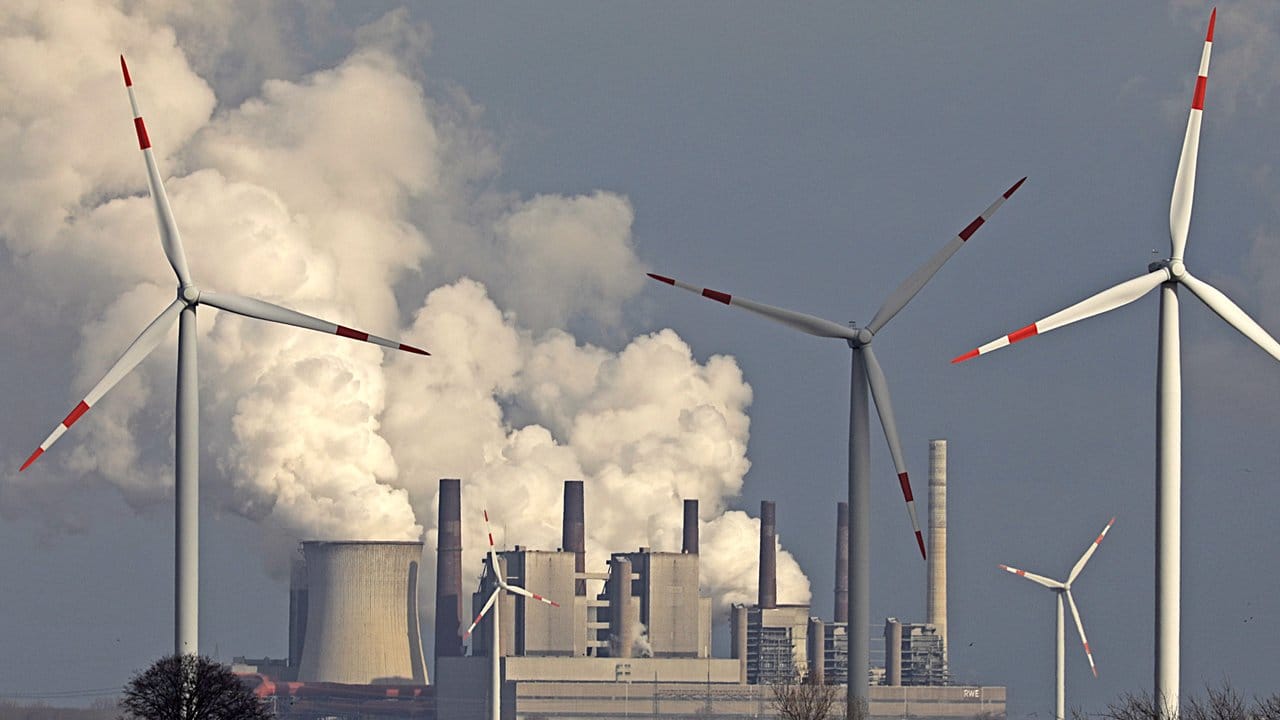 Alte und neue Technik: Rauch und Dampf steigen aus einem Braunkohlekraftwerk, im Vordergrund stehen Windräder.