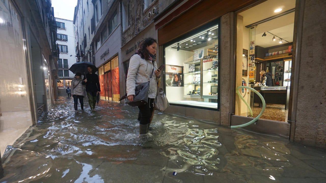Menschen waten in Gummistiefeln durch Hochwasser in einer Gasse in Venedig.