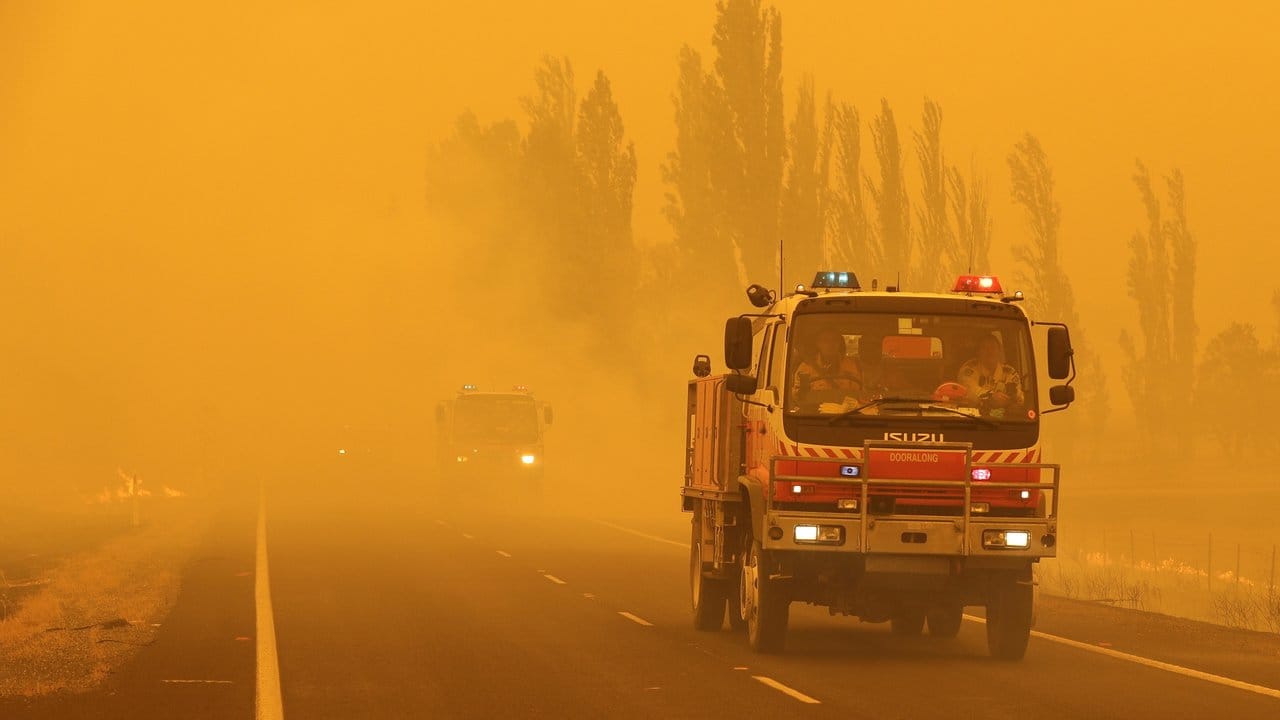 Feuerwehrfahrzeuge fahren in dichtem Rauch in der Region Canberra auf einer Straße, an deren Seiten das Gras brennt. Feuerwehrfahrzeuge fahren in dichtem Rauch in der Region Canberra auf einer Straße, an deren Seiten das Gras brennt.