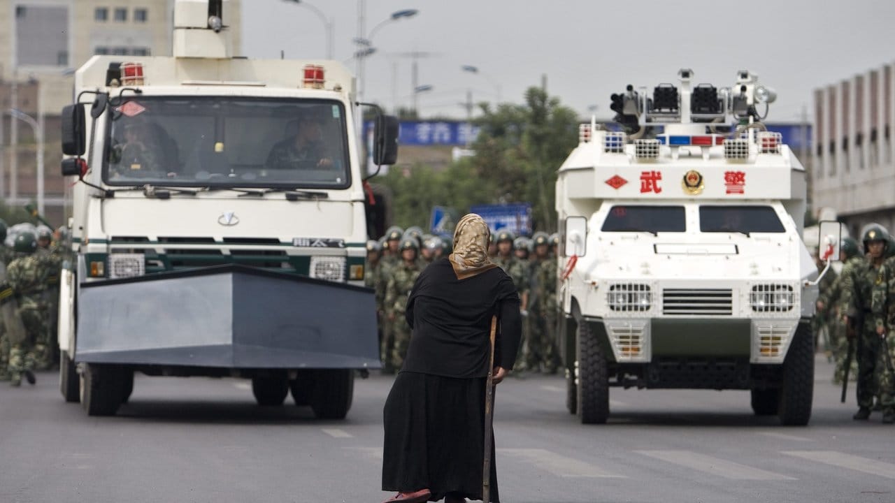 Eine uigurische Frau steht vor gepanzerten Fahrzeugen der Staatsmacht.