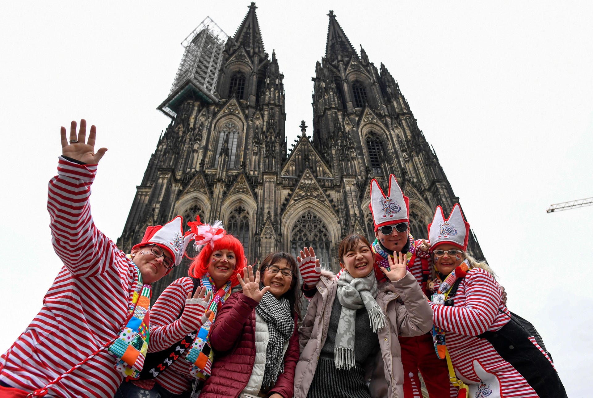 Eine Gruppe verkleideter Karnevalisten nimmt vor dem Kölner Dom zwei japanische Touristinnen in den Arm.