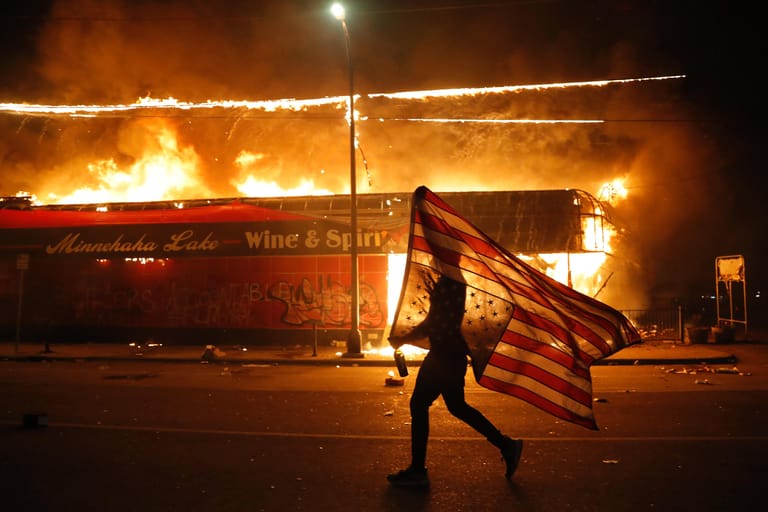 Ein Demonstrant trägt US-Flagge, während er an brennenden Gebäuden entlang läuft.