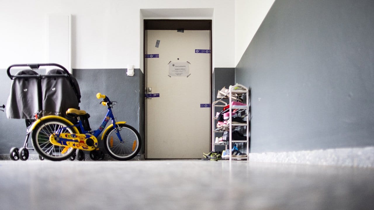 Siegel der Polizei hängen an einer Wohnungstür in einem Mehrfamilienhaus, davor stehen ein Kinderwagen, ein Kinderrad und ein Schuhregal.