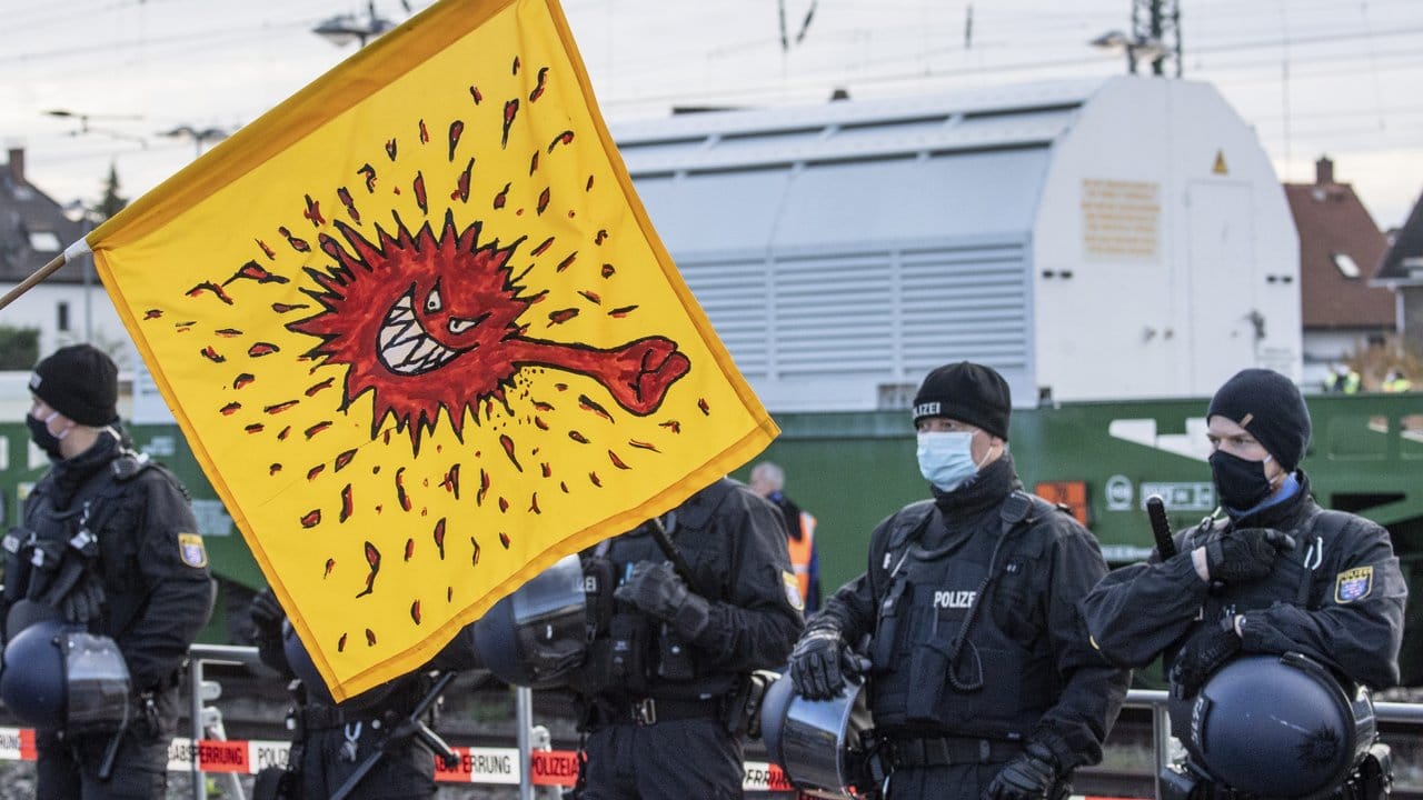 Mit einer Coronavirus-Karikatur auf einer gelben Fahne protestieren Anti-Atomkraft-Gegner neben Polizisten auf dem Bahnhof von Biblis gegen den Transport der insgesamt sechs Castor-Behälter, die auf einem Spezialzug eingetroffen sind.