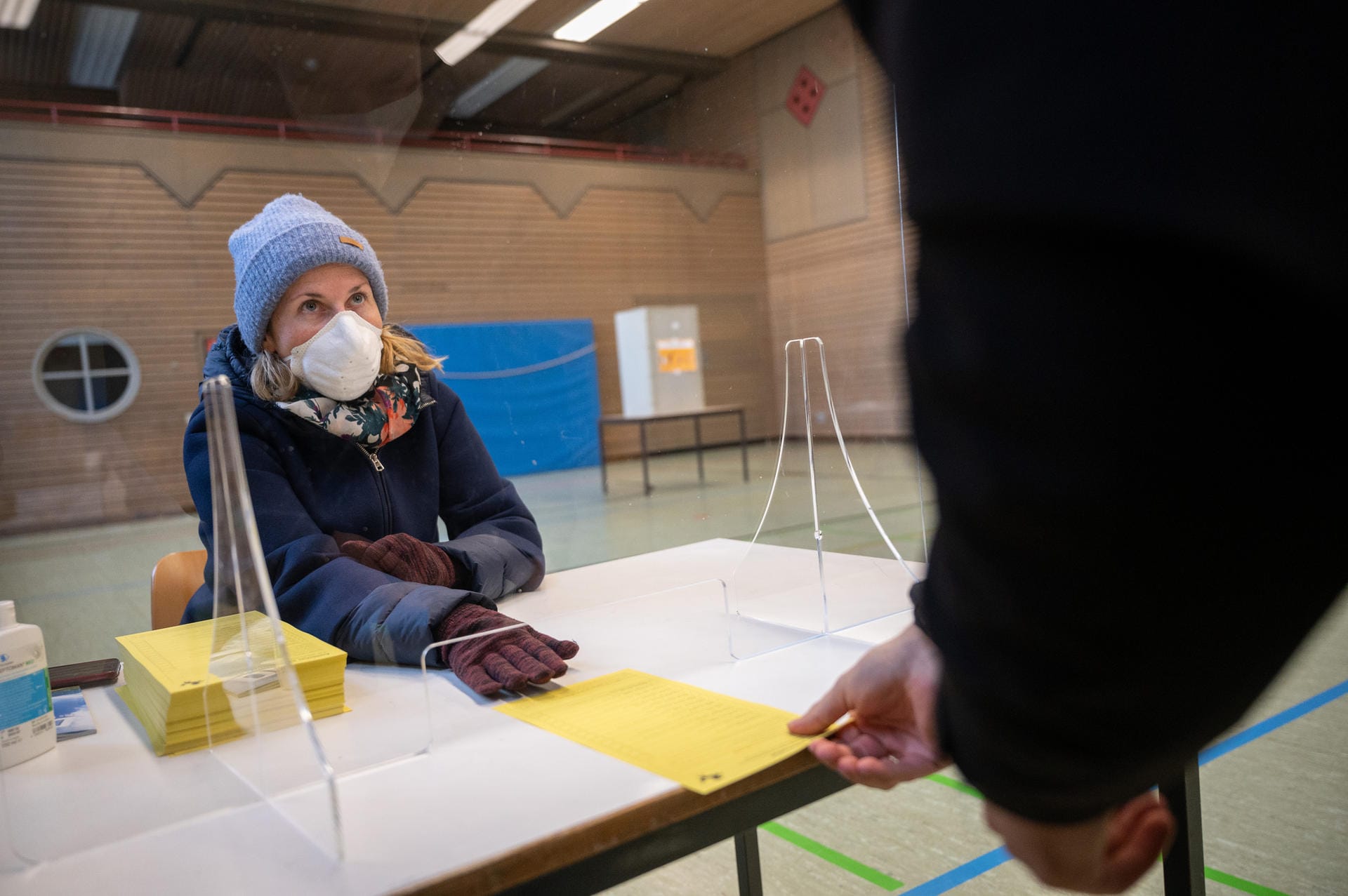 Eine Wahlhelferin mit Mundschutz sitzt hinter einer Plexiglasscheibe: Sie gibt einem Wähler einen Stimmzettel.