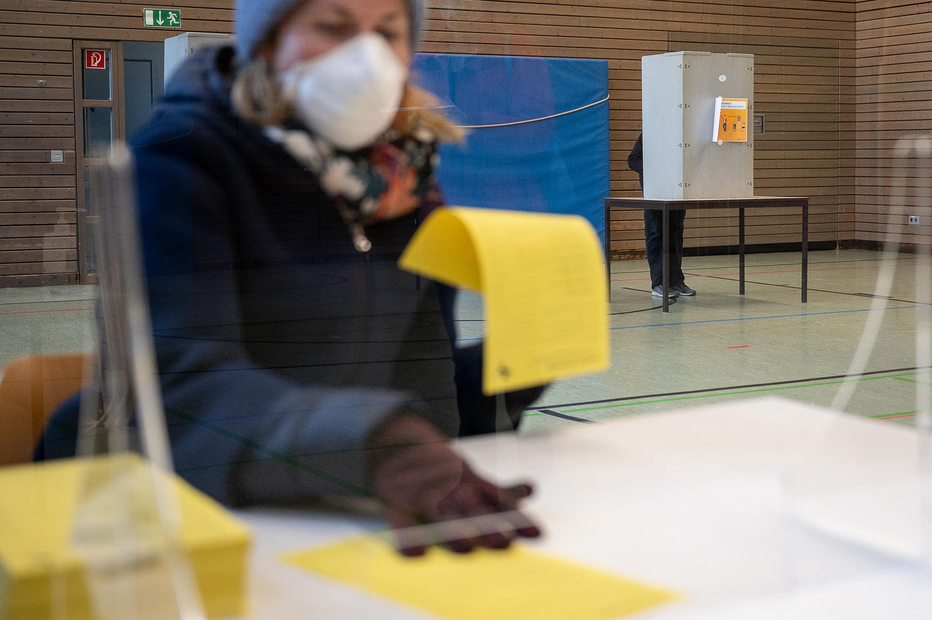 Eine Wahlhelferin mit Mund-Nasen-Schutz: Im Hintergrund steht ein Mann in einer Wahlbox.