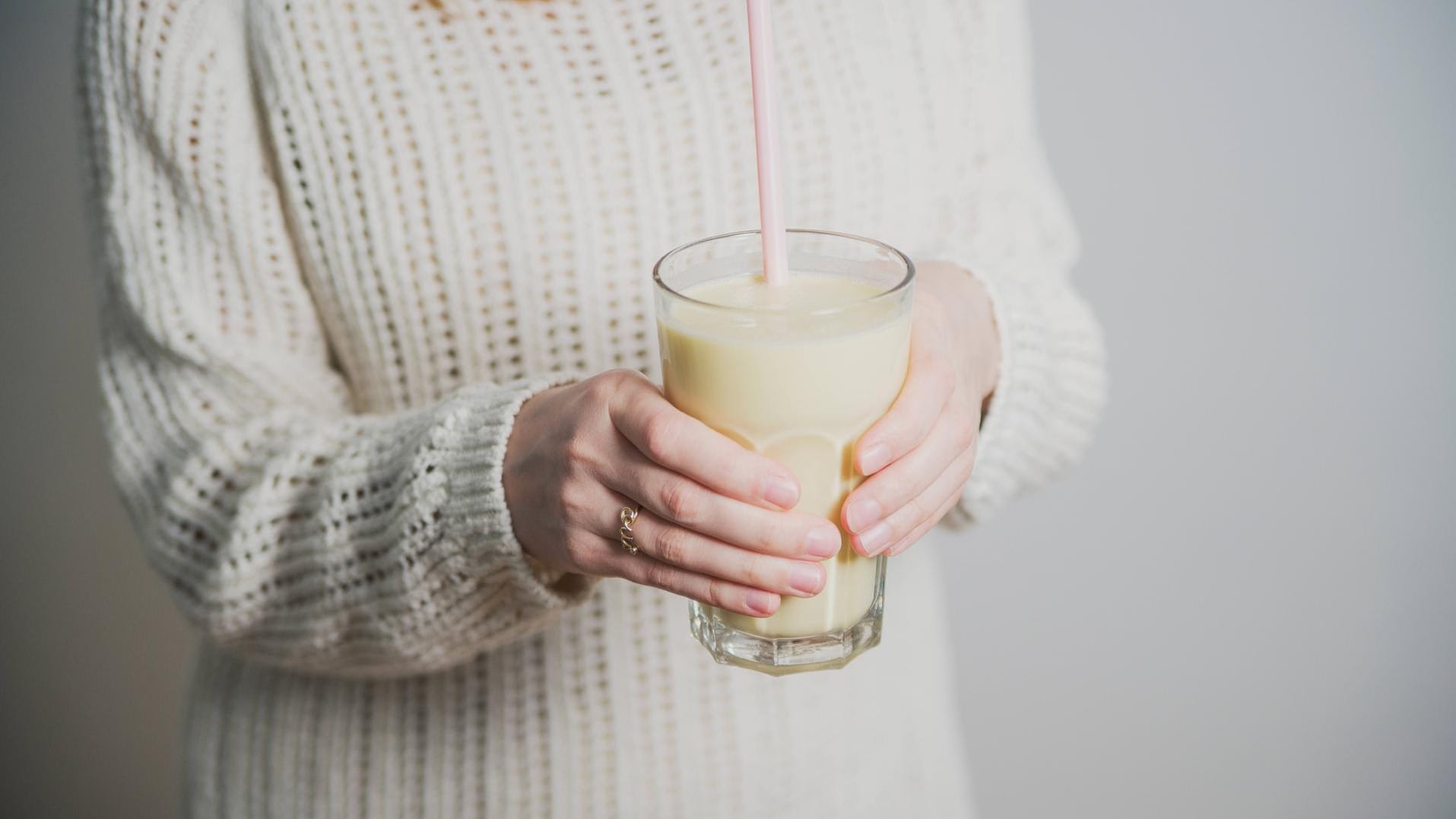 Junge Frau in weißem Pullover hält ein Glas Buttermilch mit Strohhalm.