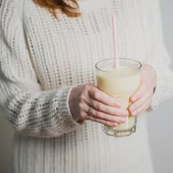 Junge Frau in weißem Pullover hält ein Glas Buttermilch mit Strohhalm.