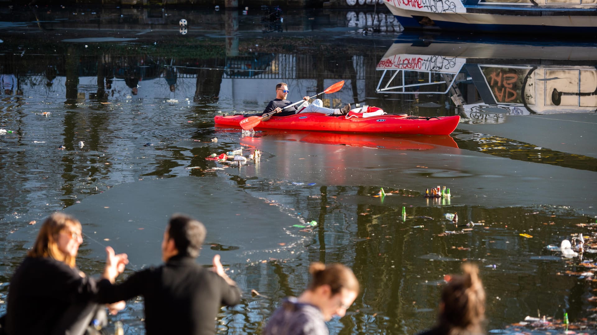 Berlin: Ein Mann fährt mit einem Kajak auf dem Landwehrkanal in Kreuzberg, während Menschen am Ufer die Sonne genießen.