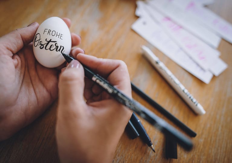 Auf ein weißes Ei wird mit einem Stift "frohe Ostern" geschrieben.