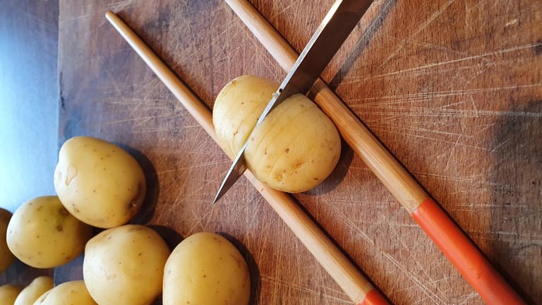Platzieren Sie die Kartoffeln einzeln zwischen zwei Essstäbchen und schneiden Sie sie fächerförmig ein – es sollten zwei bis drei Millimeter dünne Scheiben entstehen. Schneiden Sie die Kartoffeln aber nicht ganz durch, die Stäbchen bremsen deshalb den Schnitt ab.