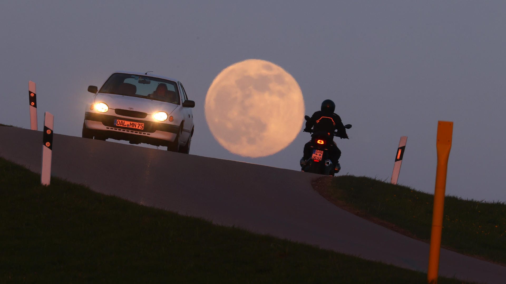 Bayern, Bernbeuren: Ein Motorrad und ein Auto fahren vor dem aufgehenden Vollmond über eine Hügelkuppe.