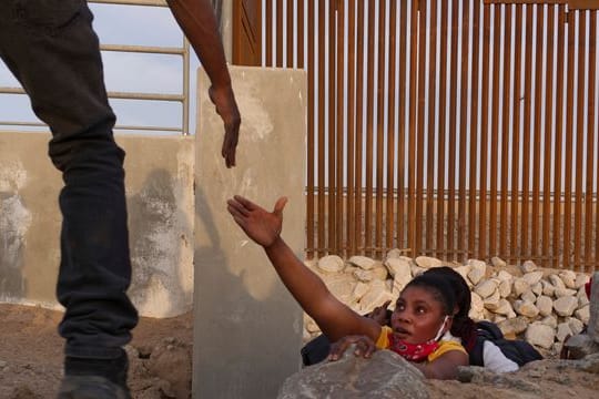 Eine Frau streckt die Hand nach Hilfe aus, während sie aus einem felsigen Kanal neben einer Lücke in der US-Grenzmauer an der mexikanischen Grenze klettert.